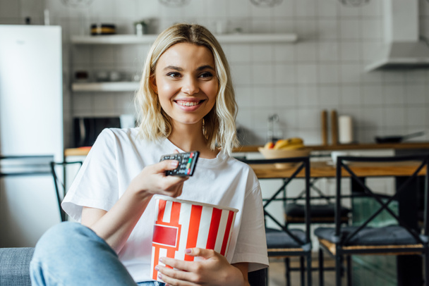 Selective focus of smiling girl holding bucket with popcorn and remote controller on couch  - Fotografie, Obrázek