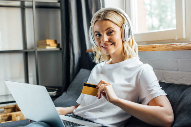 Menina atraente em fones de ouvido sorrindo para a câmera enquanto segurando cartão de crédito e laptop na sala de estar
 - Foto, Imagem