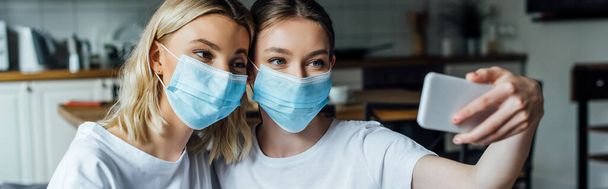 Panoramic shot of sisters in medical masks taking selfie with smartphone at home  - Photo, Image