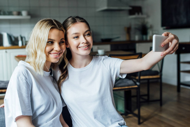 Beautiful smiling sisters taking selfie with smartphone at home  - Valokuva, kuva