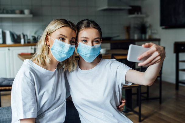 Αδελφές με ιατρικές μάσκες που βγάζουν selfie με smartphone στο σπίτι  - Φωτογραφία, εικόνα