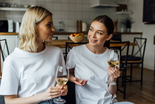 Smiling sisters talking while holding glasses of wine at home  - Fotografie, Obrázek