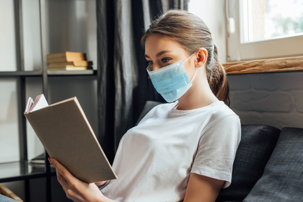 Young woman in medical mask reading book on sofa  - Photo, Image