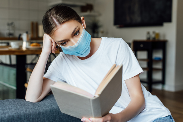 Enfoque selectivo de la mujer joven en el libro de lectura de máscara médica en la sala de estar
 - Foto, imagen