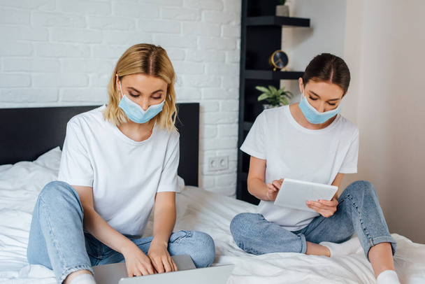 Sisters in medical masks using laptop and digital tablet on bed  - Photo, Image