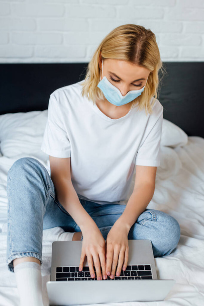 Selective focus of blonde girl in medical mask using laptop while sitting on bed  - Фото, изображение