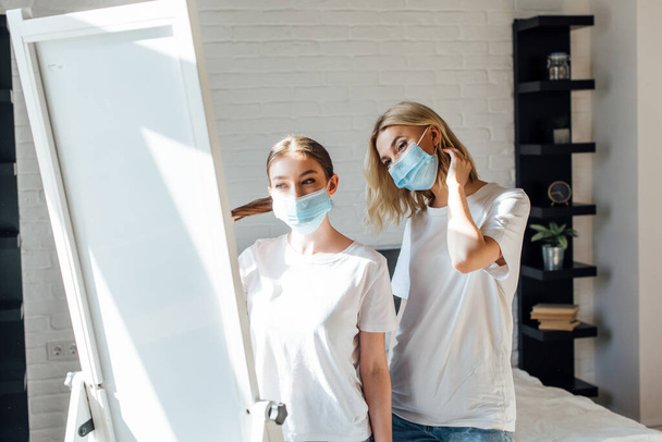 Sisters in medical masks standing near mirror in bedroom  - Photo, Image