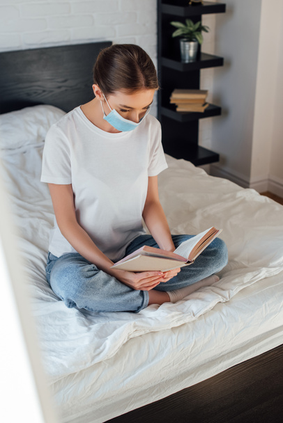 Selective focus of young woman in medical mask reading book on bed  - Photo, Image