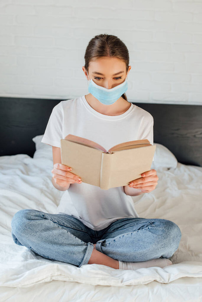 Junge Frau in medizinischer Maske liest Buch, während sie mit überkreuzten Beinen auf dem Bett sitzt  - Foto, Bild