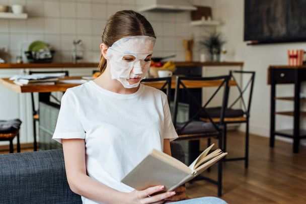 Selective focus of young woman in face mask reading book at home  - Foto, afbeelding