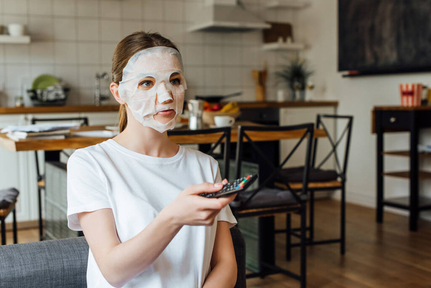 Νεαρή γυναίκα με μάσκα προσώπου κρατώντας τηλεχειριστήριο στο σπίτι  - Φωτογραφία, εικόνα