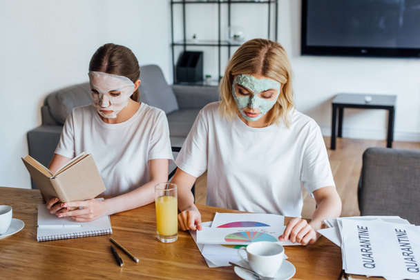 Sisters in face masks reading book and working with papers at table  - Фото, изображение