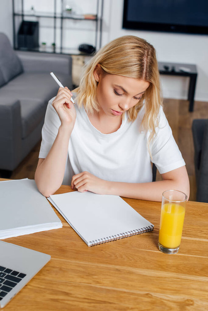 Blondes Mädchen hält Stift neben Notizbuch, Laptop und einem Glas Orangensaft zu Hause  - Foto, Bild