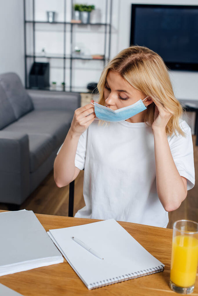 Selective focus of blonde woman putting on medical mask near papers, notebook and glass of orange juice on table  - Foto, immagini
