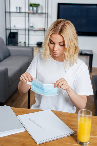 Selective focus of blonde woman holding medical mask near orange juice and papers on table  - Foto, afbeelding