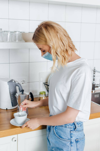 Vista lateral da mulher loira em máscara médica misturando café na xícara na cozinha
  - Foto, Imagem