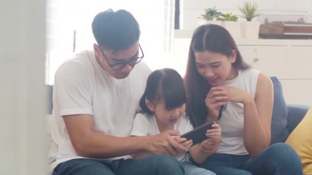 Happy Asian family dad, mom and daughter playing funny game online on smartphone sitting sofa in room at house. Self-isolation, stay at home, social distancing, quarantine for coronavirus prevention. - Video