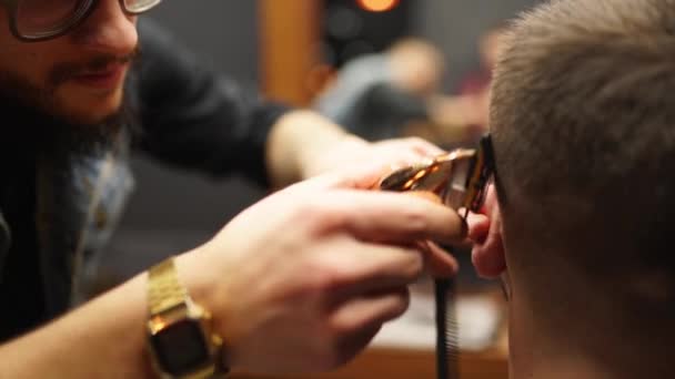 Trendy barber cuts bearded mans hair with a clipper in barbershop. Mens hairstyling and hair cutting in salon. Grooming the hair with trimmer. Hairdresser doing haircut in retro hair salon. - Filmati, video