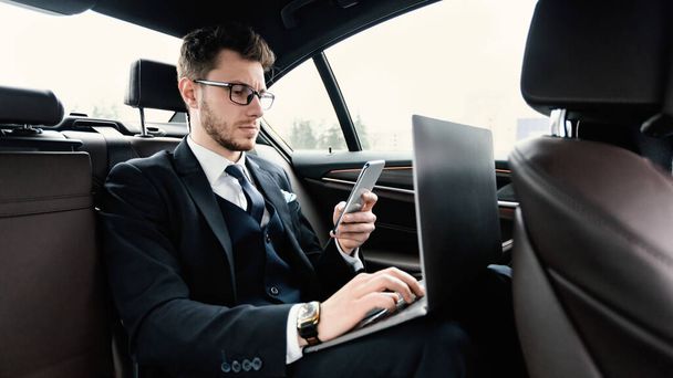 Lüks bir arabada oturan bir iş adamı telefon ve bilgisayar kullanıyor. - Fotoğraf, Görsel
