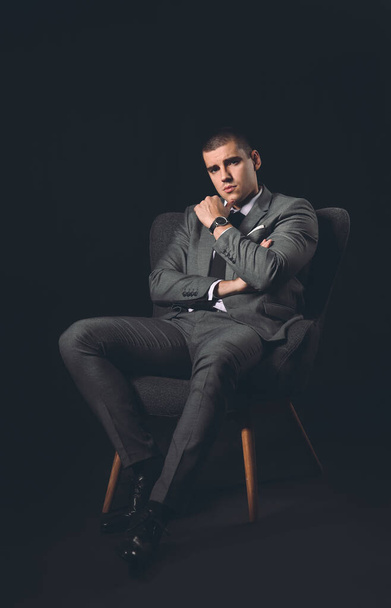 Handsome young man in elegant suit sitting in armchair on dark background - Foto, imagen