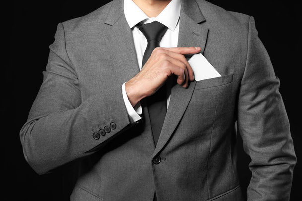 Schöner junger Mann im eleganten Anzug zieht Visitenkarte vor dunklem Hintergrund aus der Tasche - Foto, Bild