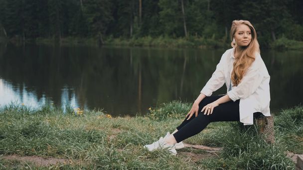 Beautiful young blonde woman natural appearance, long hair, beautiful face, sitting in a park near the lake. Side view - Фото, зображення