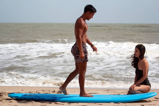 Jeune homme beau debout sur longboard, enseignant belle dame pour le modèle de base du surf, sur la plage  - Photo, image