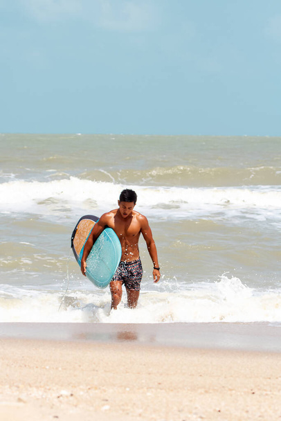 Joven hombre guapo mojado por todo el cuerpo sosteniendo tabla de surf con la mano derecha, caminando en la playa, mostrar el cuerpo adecuado y firme, modelo de posición, luz borrosa alrededor. - Foto, imagen