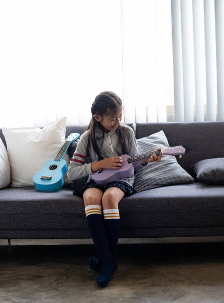 The little girl sitting on sofa,playing ukulele with happy feeling,blurry light around - Foto, Imagem