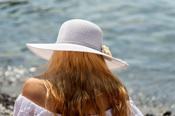 Close-up of a young blonde woman with a white straw hat and white dress on the beach with blurred sea in the background. Vernazza, Liguria, Italy - Photo, image