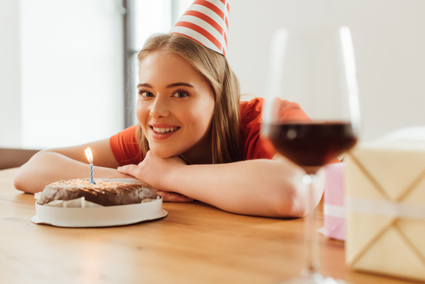 selective focus of happy girl in party cap looking at camera near birthday cake, presents and wine glass on table - Fotografie, Obrázek