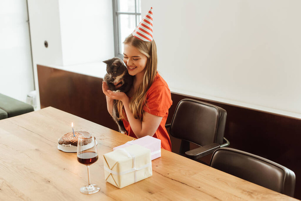 chica feliz en partido gorra celebración en brazos gato y mirando pastel de cumpleaños cerca de regalos y vino tinto en vidrio
 - Foto, Imagen