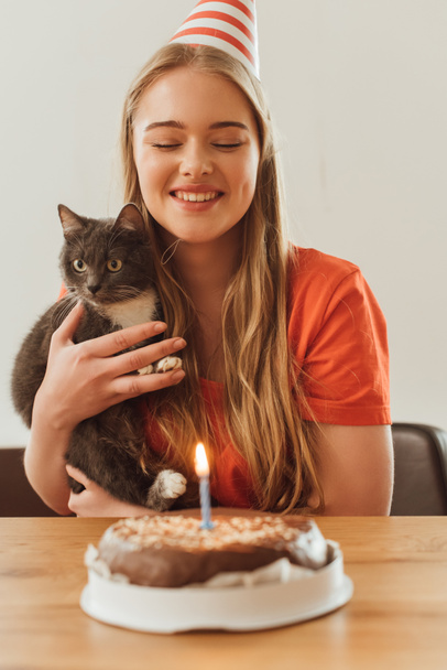 selective focus of happy girl looking at burning candle on birthday cake and holding in arms cute cat  - Photo, Image