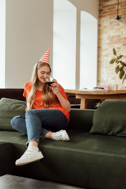 happy young woman in party cap sitting on sofa and drinking red wine  - Foto, imagen