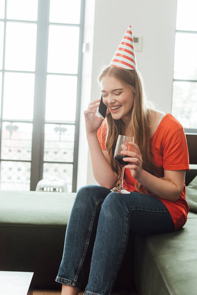 smiling girl in party cap holding glass of red wine and talking on smartphone at home  - Photo, Image