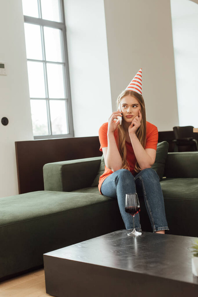 selective focus of displeased girl in party cap sitting on sofa and talking on smartphone at home  - Foto, imagen