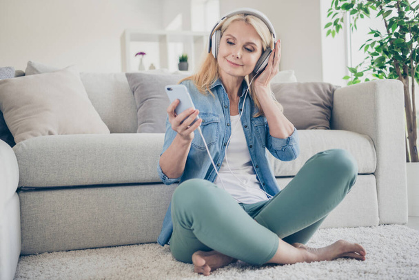 Photo of funny feel young middle aged domestic lady sit comfy floor couch browsing telephone listen music cool earphones remote work stay home quarantine weekend living room indoors - Foto, Bild