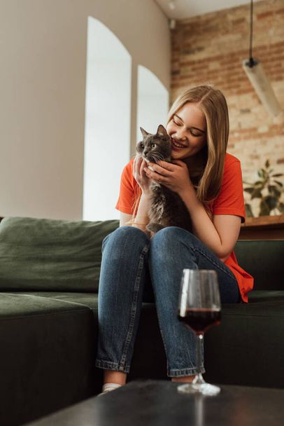 Seçici odak noktası, kırmızı şarap kadehinin yanında sevimli bir kedi tutan mutlu genç kadın.  - Fotoğraf, Görsel