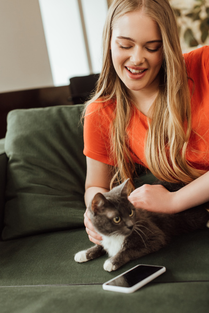 Kanepede boş ekranlı akıllı telefonun yanındaki sevimli kediye dokunan neşeli genç kadın. - Fotoğraf, Görsel