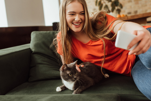 selektywne skupienie się na uśmiechniętej młodej kobiecie robiącej selfie z kotem  - Zdjęcie, obraz