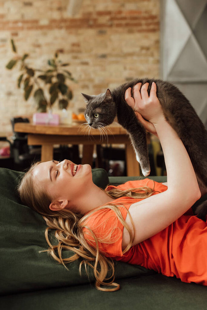 Kanepede uzanmış, kollarında sevimli bir kedi tutan neşeli bir kadın.  - Fotoğraf, Görsel