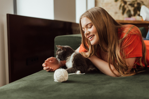 cheerful girl playing cute cat near ball of yarn on sofa - Fotoğraf, Görsel