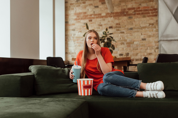 attractive girl eating popcorn, holding disposable cup and watching movie in living room  - Foto, afbeelding