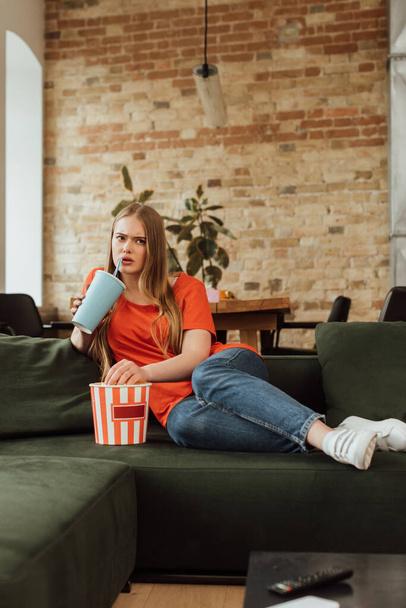 selective focus of woman drinking soda near popcorn bucket and watching movie in living room  - Foto, imagen