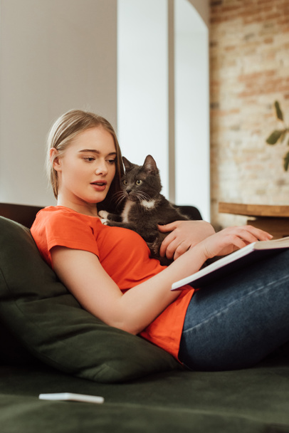 selective focus of beautiful girl holding cat and reading book in living room  - Foto, afbeelding