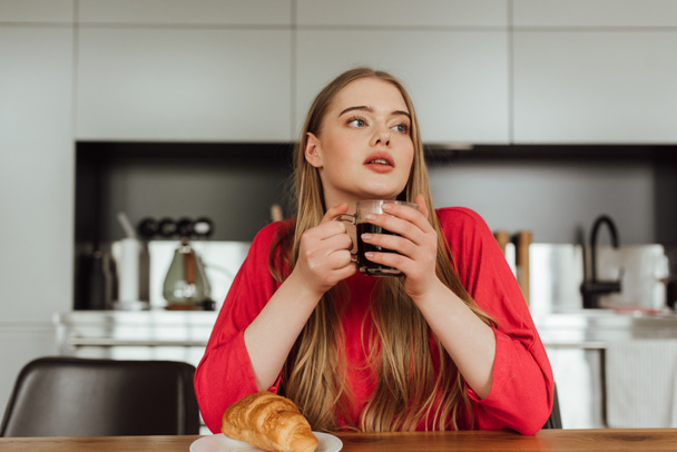 young woman holding cup of coffee near tasty croissant on table  - Photo, Image