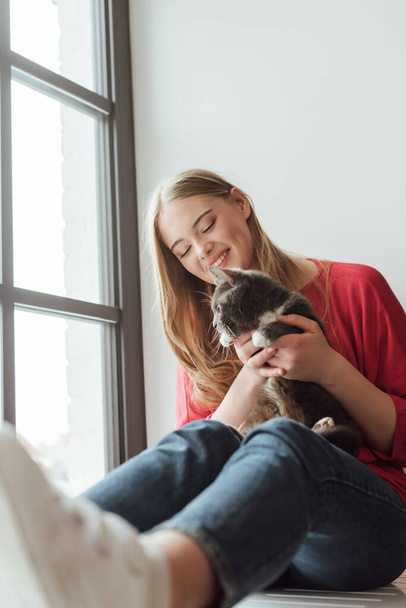 selective focus of positive woman holding cute cat while sitting on window sill  - Foto, imagen