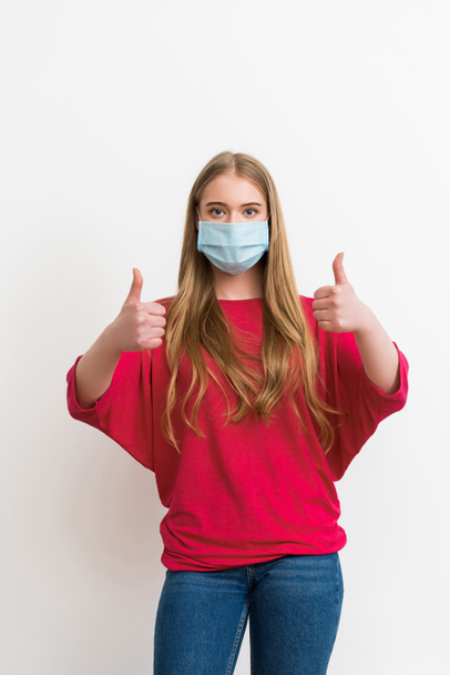молодая женщина в медицинской маске показывает большие пальцы вверх изолированы на белом
  - Фото, изображение