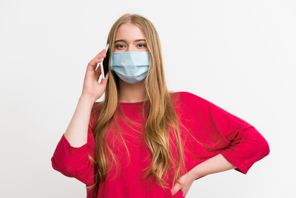 young woman in medical mask talking on smartphone and standing with hand on hip isolated on white  - Fotografie, Obrázek