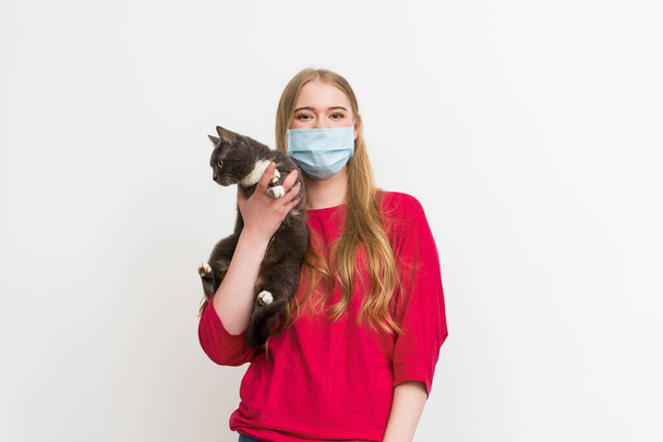 junge Frau in medizinischer Maske hält süße Katze im Arm und schaut in die Kamera isoliert auf weiß  - Foto, Bild